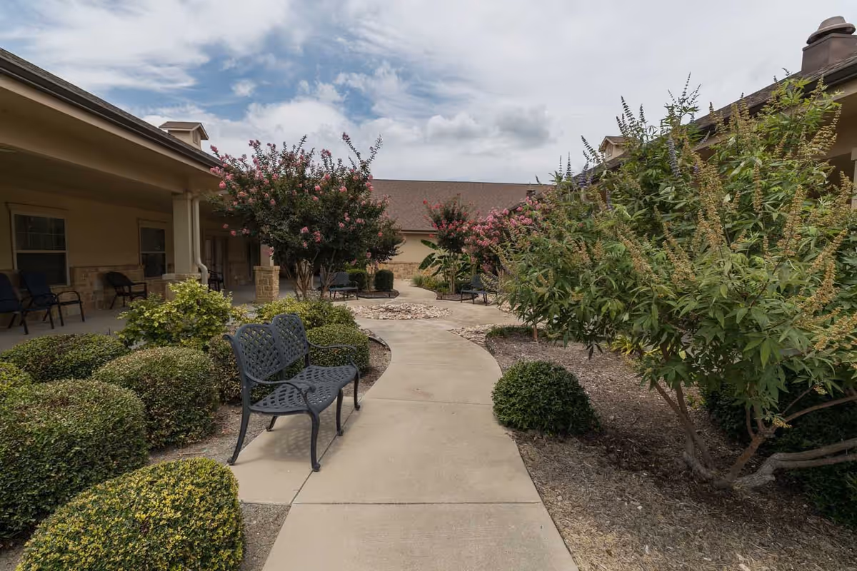 Outdoor courtyard area with a concrete walkway lined by trimmed bushes and flowering trees. Metal benches are placed along the path, and the buildings with covered walkways and windows surround the courtyard under a partly cloudy sky.