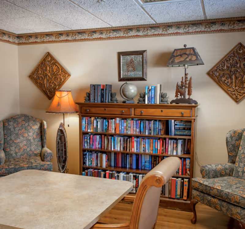 Cozy room with a wooden bookshelf filled with books, two floral upholstered armchairs, a table with a light-colored surface, a standing lamp with a warm light, and decorative wall art including a framed picture and carved wooden panels.