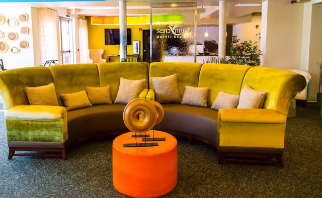 A bright senior living lobby with a curved chartreuse sofa, an orange ottoman topped by a decorative sculpture, and a glass panel showing the facility name.