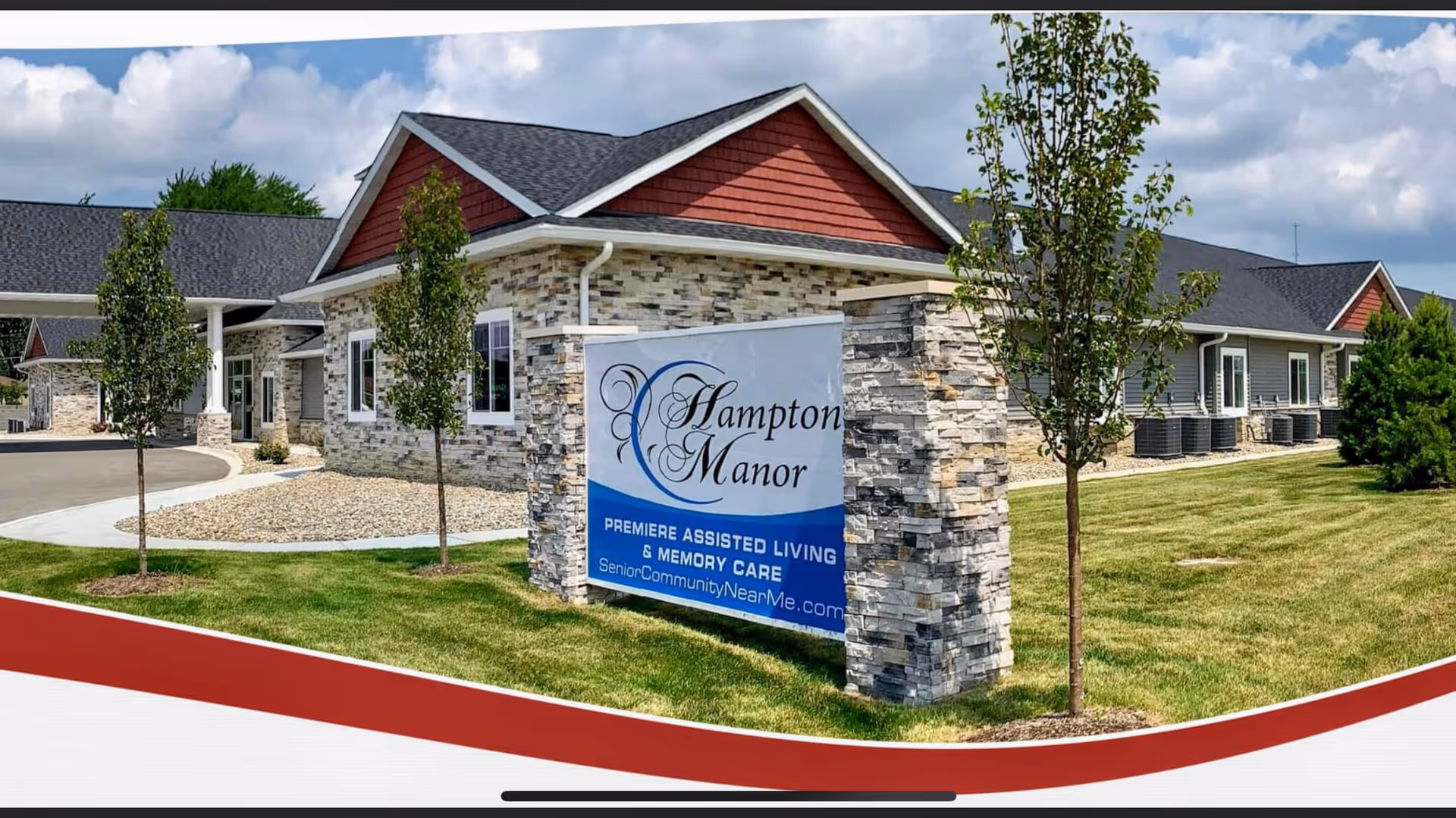Exterior view of Hampton Manor, a single-story assisted living and memory care facility with stone and siding walls, a dark shingled roof, and a landscaped lawn with small trees. A large stone sign in front displays the facility name and services.
