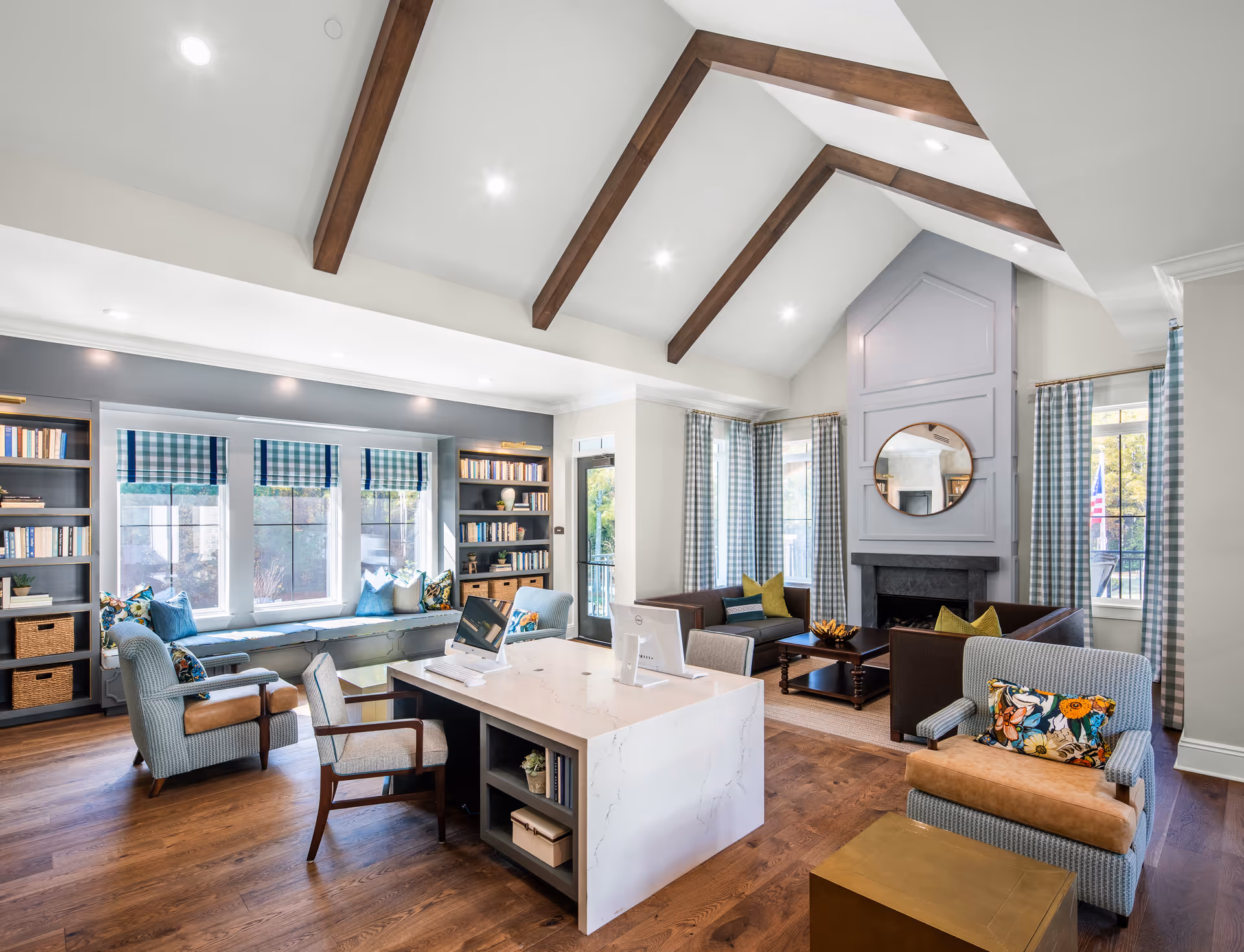 Sunlit communal lounge with vaulted beamed ceiling, fireplace, built-in bookshelves, window seat, desk, and multiple seating areas.