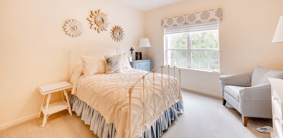 A bright bedroom with a white metal bed frame, cream-colored bedding, and decorative pillows. There is a small white side table next to the bed, a blue dresser with a lamp and decorative items, and a comfortable gray armchair near a large window with a patterned valance. The walls are light beige and decorated with three sunburst mirrors.