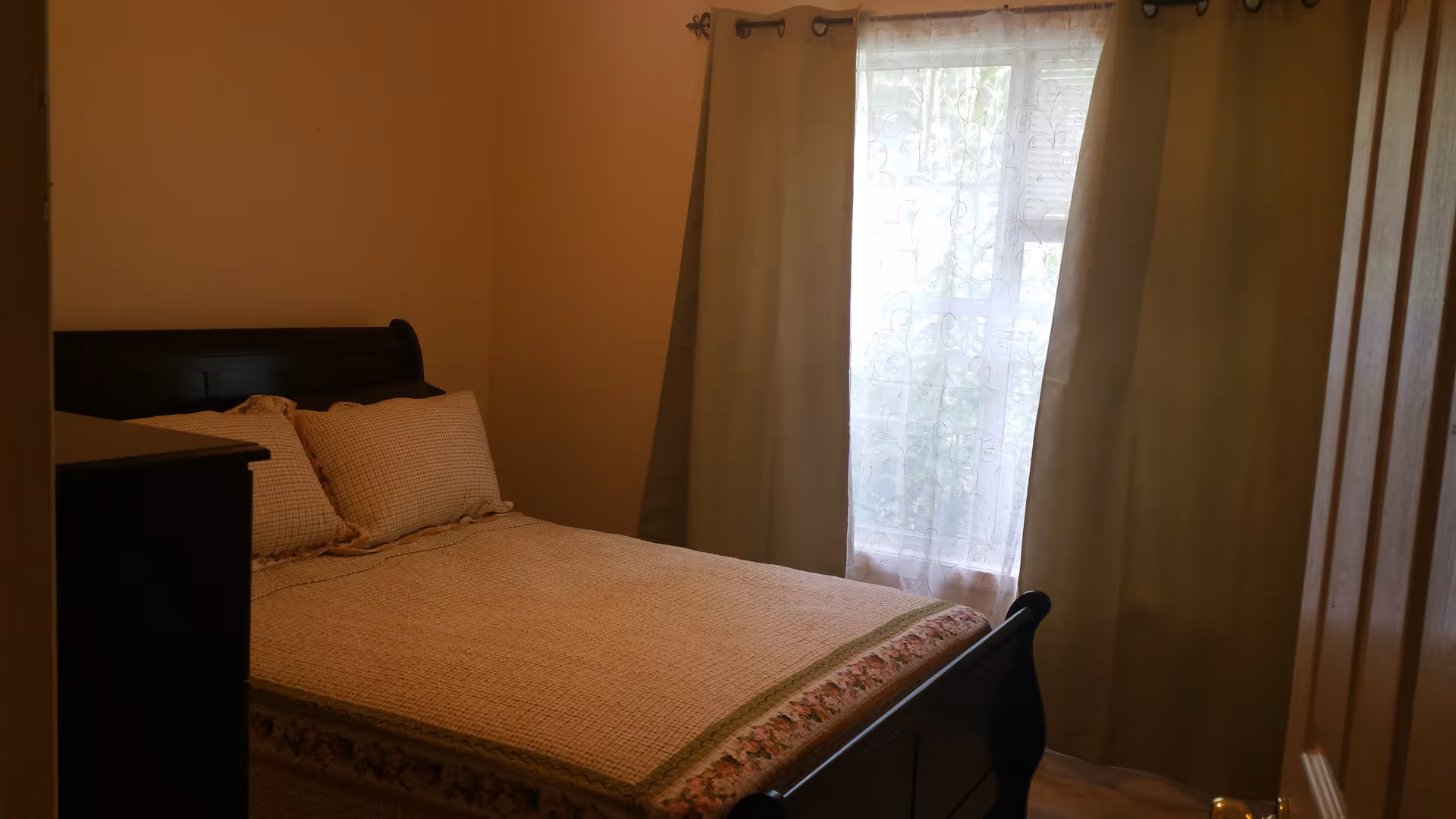 Small bedroom with a made bed, dark wood headboard and dresser, and a window with curtains letting in daylight.