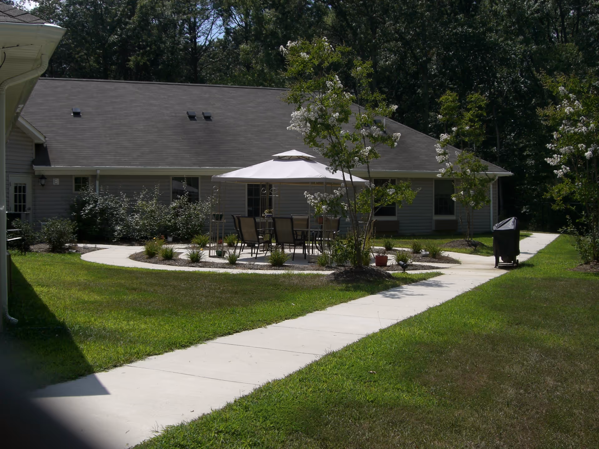 Outdoor patio area at Fenwick Landing Senior Care Community with a white gazebo covering a table and several chairs, surrounded by small trees and plants. A concrete walkway curves around the patio, leading to a gray building with a shingled roof in the background, and a grill covered with a black cover is visible on the right side.