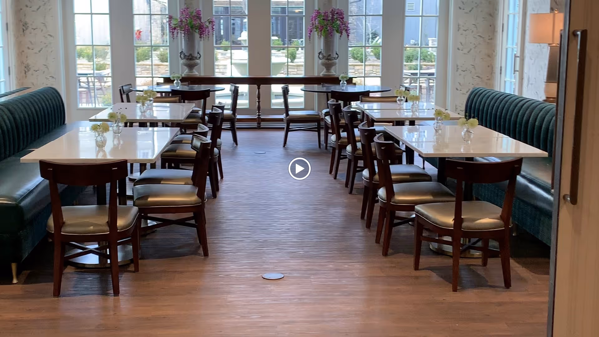 Sunlit dining room with rows of tables and chairs flanked by green banquette seating and vases of flowers in front of large windows.