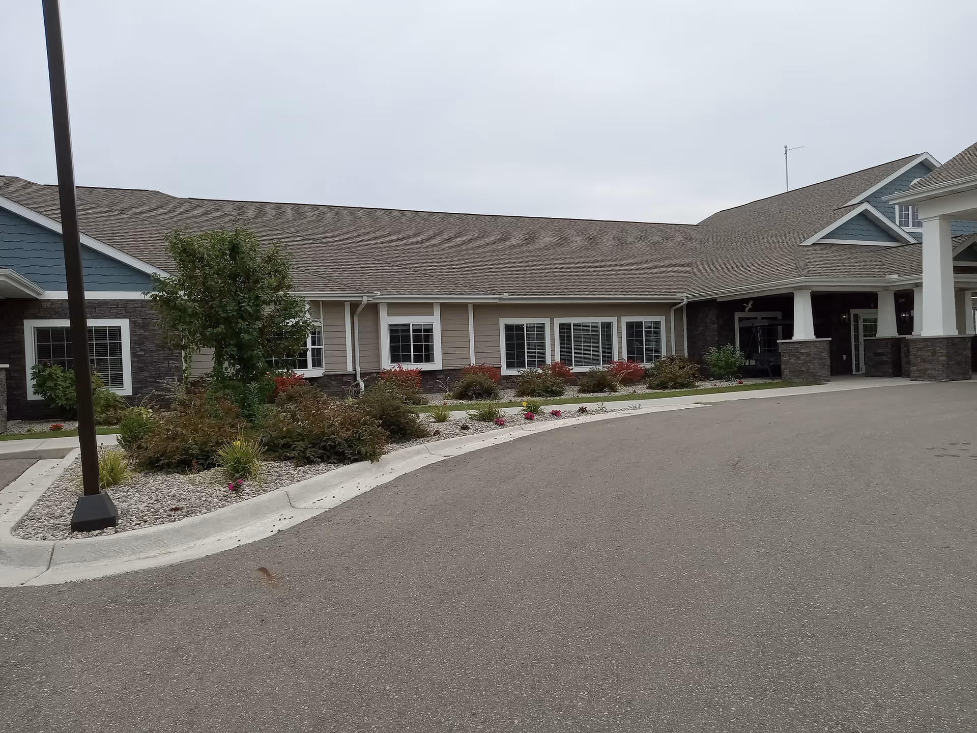 Front entrance and circular driveway of a single-story assisted living facility with landscaped beds and large windows.