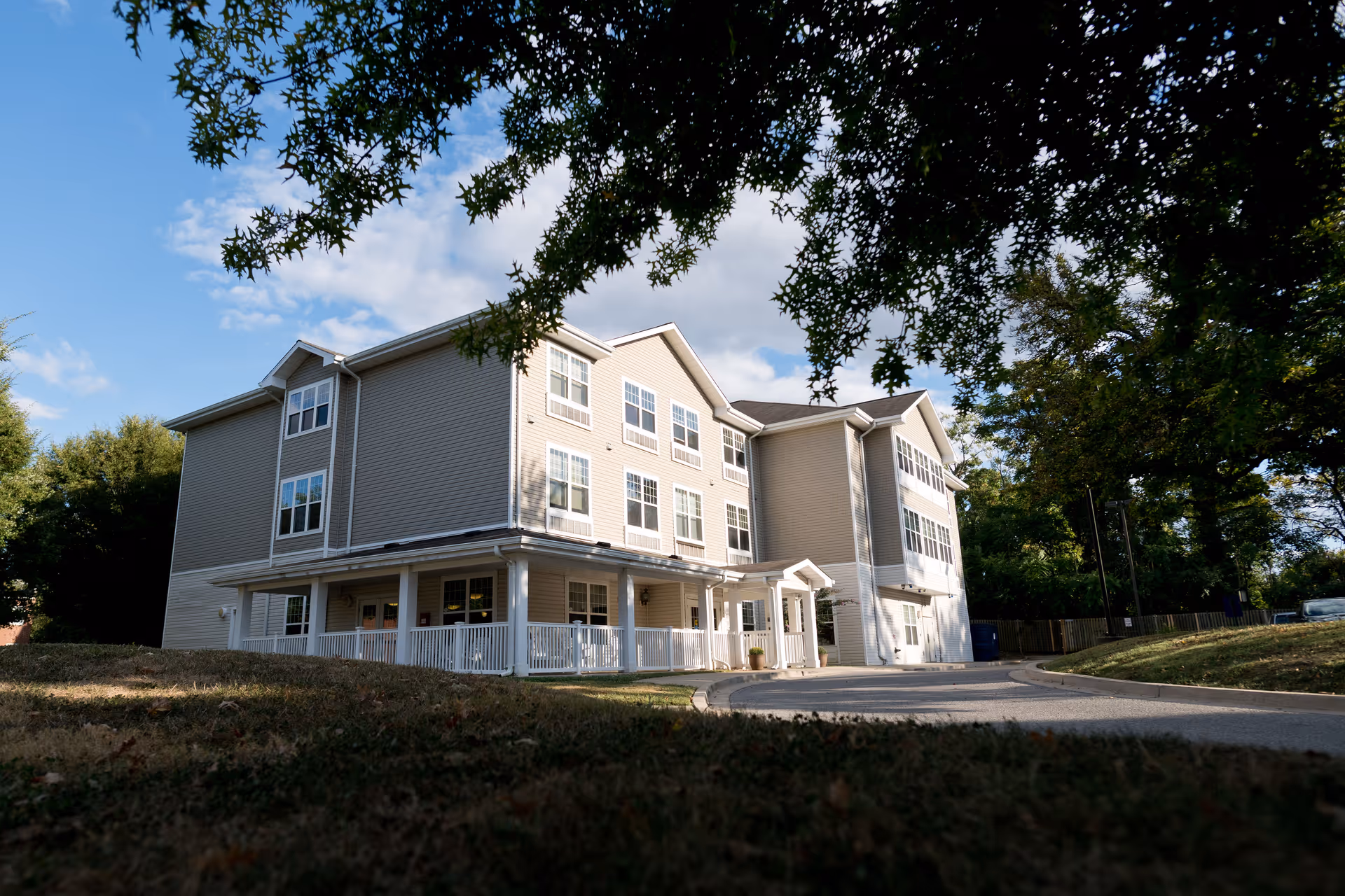 Exterior view of a three-story beige senior living facility building with white trim and multiple windows, surrounded by trees and a driveway, under a partly cloudy blue sky.