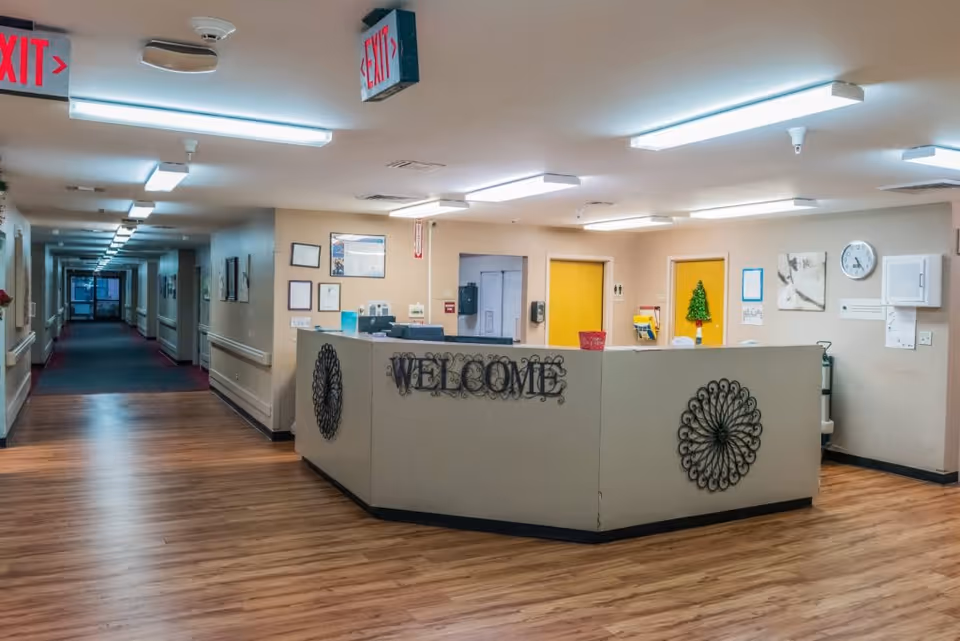 Reception area inside Chelsea Gardens Skilled Nursing & Rehab Center with a welcome desk decorated with metal wall art and a small Christmas tree. The area has wood flooring, fluorescent ceiling lights, and a hallway extending to the left with exit signs visible.