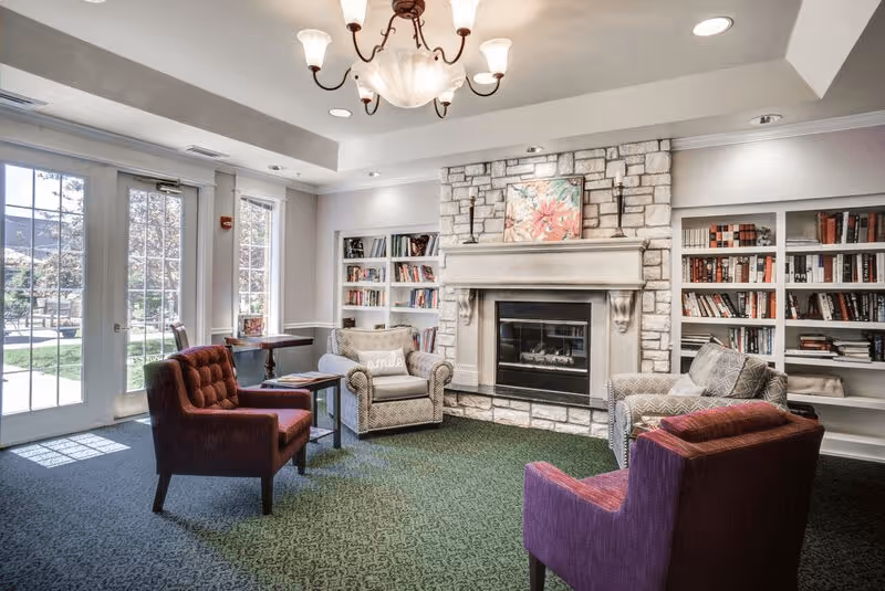 A cozy living room area with a stone fireplace in the center, flanked by built-in bookshelves filled with books. Four upholstered chairs, two in a purple fabric and two in a patterned beige fabric, are arranged around the fireplace. A small side table is placed between two chairs. Large windows and glass doors on the left allow natural light to fill the room, showing an outdoor patio area.
