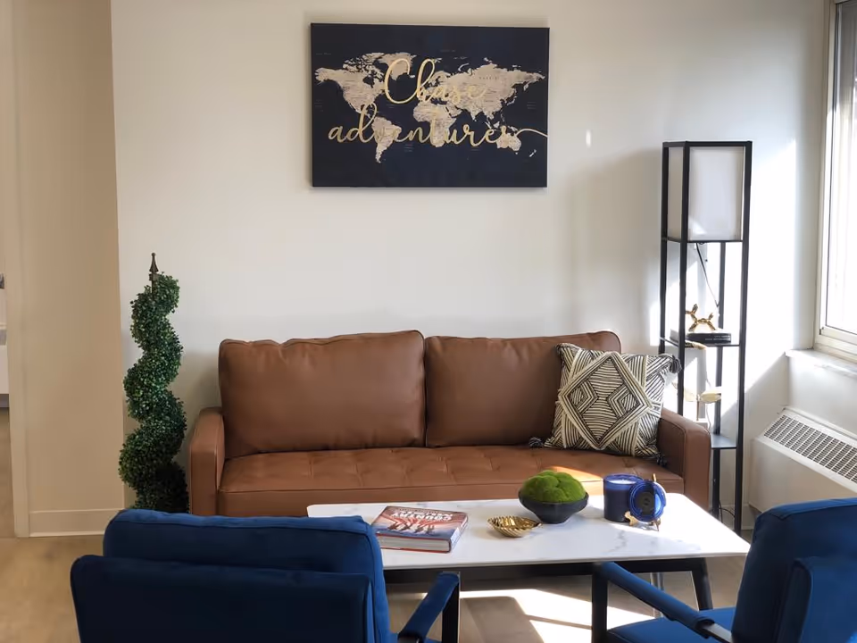 A cozy living room area with a brown leather sofa adorned with a patterned cushion, a white marble coffee table holding a book, a small green plant, a blue candle, and a decorative item. Two blue chairs face the sofa. A tall black shelving unit stands near a window letting in natural light. A wall art piece above the sofa displays a world map with the words 'Choose adventure'.