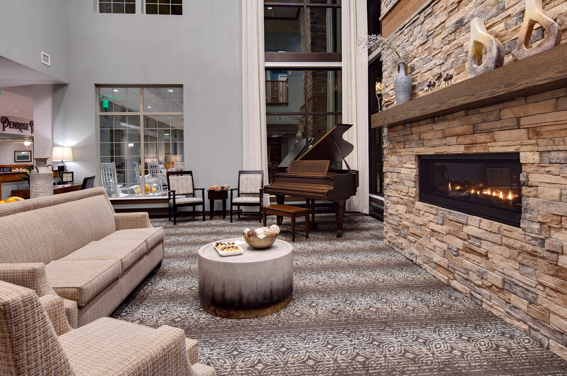 A cozy living room area in a senior living facility featuring a beige sofa and armchair, a round coffee table with snacks and decorative items, two chairs with a small table between them, a grand piano, and a stone fireplace with a lit fire. Large windows with white curtains and a patterned carpet complete the space.