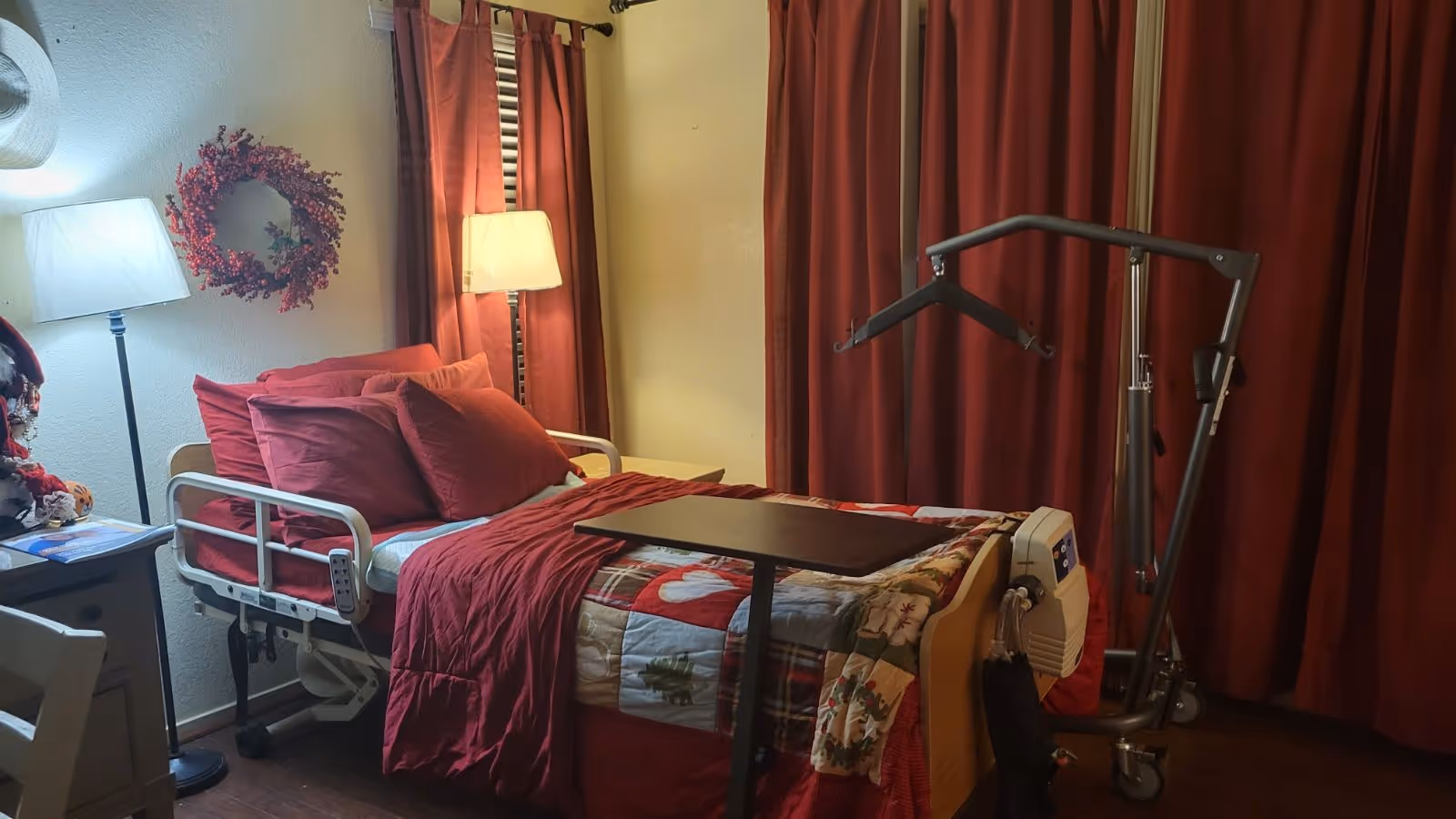 A cozy bedroom in an elder care facility featuring a hospital-style bed with red bedding and multiple pillows. There is a small overbed table on the bed, a floor lamp beside the bed, a wreath hanging on the wall, and a patient lift device next to the bed. The room has red curtains covering the windows.