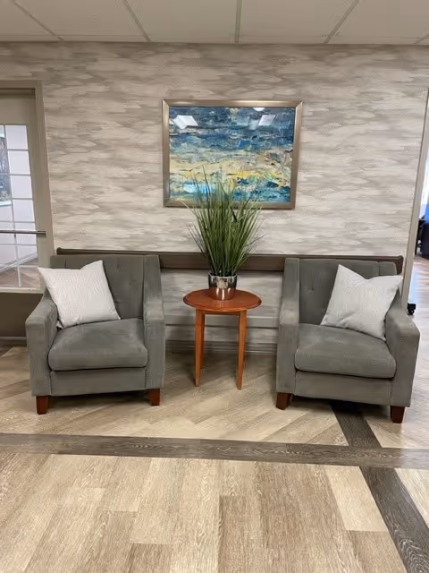 Two gray upholstered armchairs with white pillows are positioned on either side of a small round wooden table holding a potted green plant. Behind the chairs is a wall with a textured gray and white pattern and a framed abstract painting featuring blue, green, and yellow tones. The floor has a wood-like pattern with a darker border.