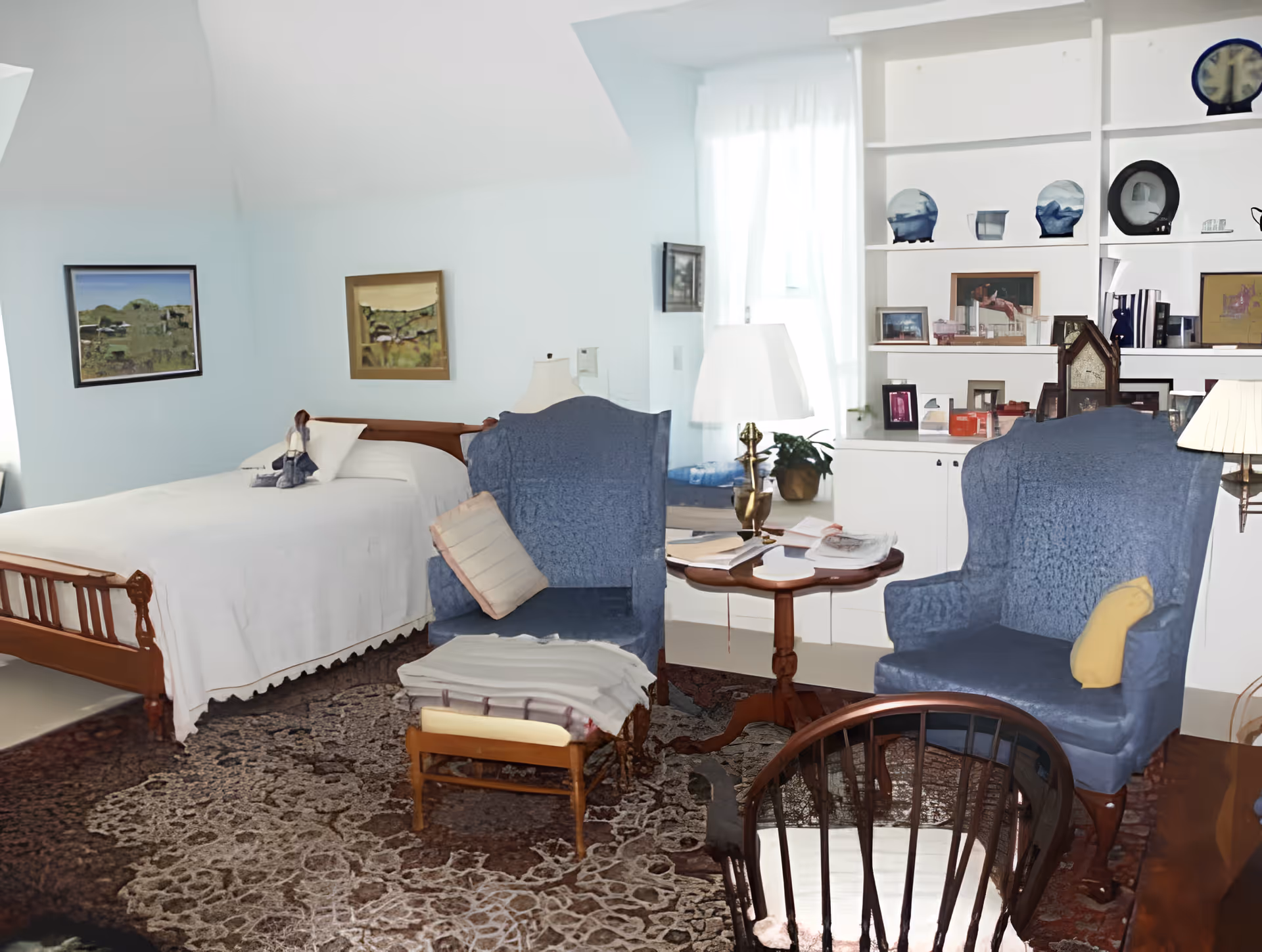 A cozy bedroom with a wooden bed covered in a white bedspread, two blue upholstered armchairs with cushions, a small wooden table with books and a lamp, a patterned rug, and built-in white shelves displaying various decorative items and framed pictures. The room has light blue walls and a window with sheer white curtains allowing natural light to enter.