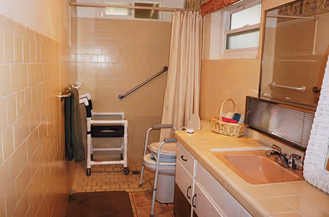 Bathroom with beige tiled walls and floor, featuring a shower area with a white shower chair and grab bar, a beige curtain, a sink with a pink basin, a mirror, a basket with towels on the countertop, and a window above the sink.