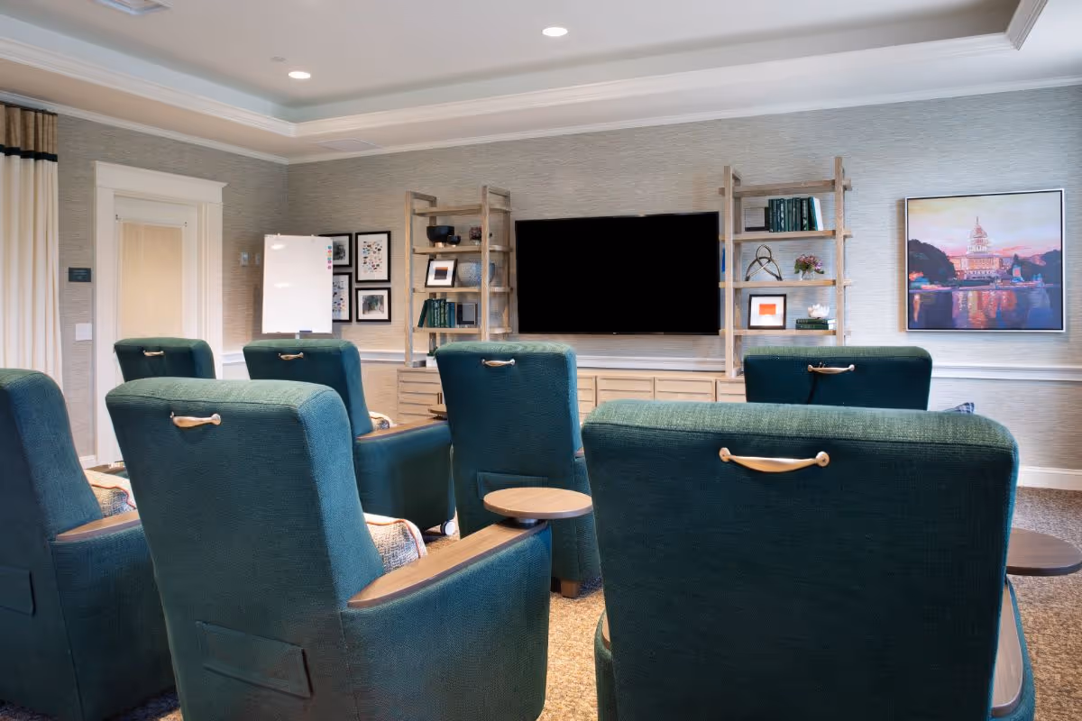 A cozy common area with six green upholstered armchairs arranged in two rows facing a large flat-screen TV mounted on a wall. The wall behind the TV has two wooden shelving units with books, framed pictures, and decorative items. There is a whiteboard on the left side and a colorful painting on the right side of the TV. The room has soft lighting and neutral-colored walls with crown molding.