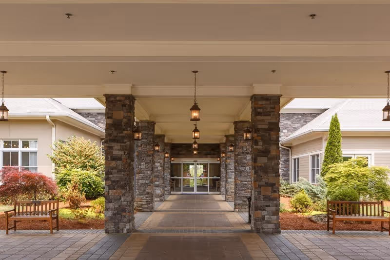 Covered entrance walkway to a building with stone pillars and hanging lantern-style lights, flanked by landscaped garden areas with bushes and two wooden benches on either side.