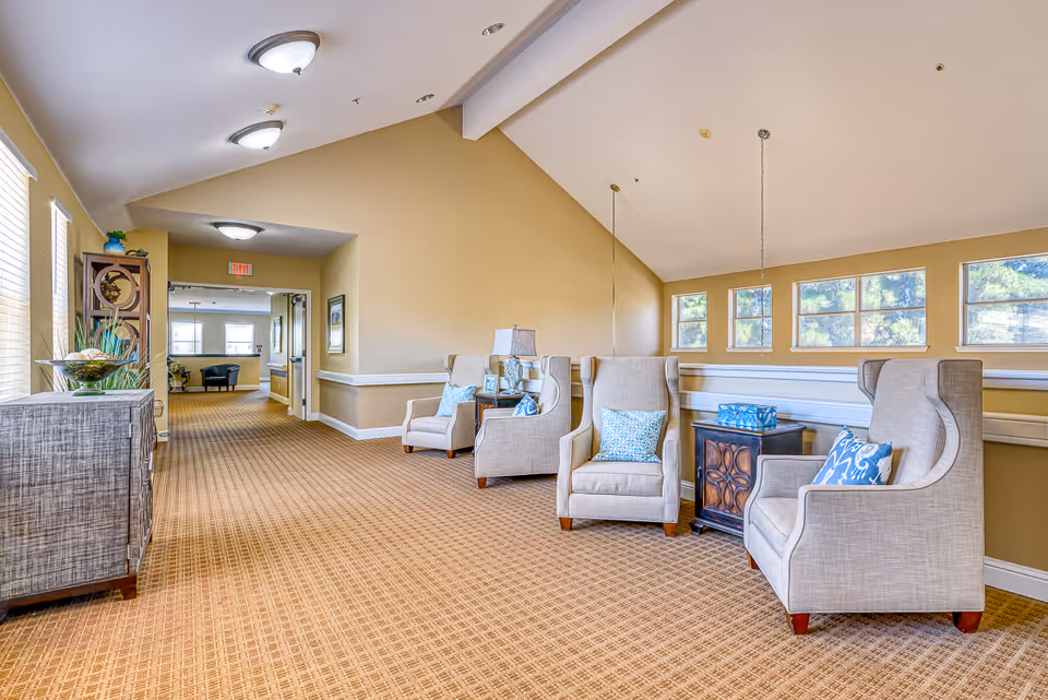 A bright and spacious hallway in a senior living facility with beige walls and carpeted floor. The hallway features several comfortable beige armchairs with blue patterned pillows arranged along the right side, accompanied by small wooden side tables. There are multiple windows letting in natural light, and decorative items including a lamp and framed photos on the side tables. The ceiling has recessed lighting fixtures, and the far end of the hallway opens into another room with additional seating.
