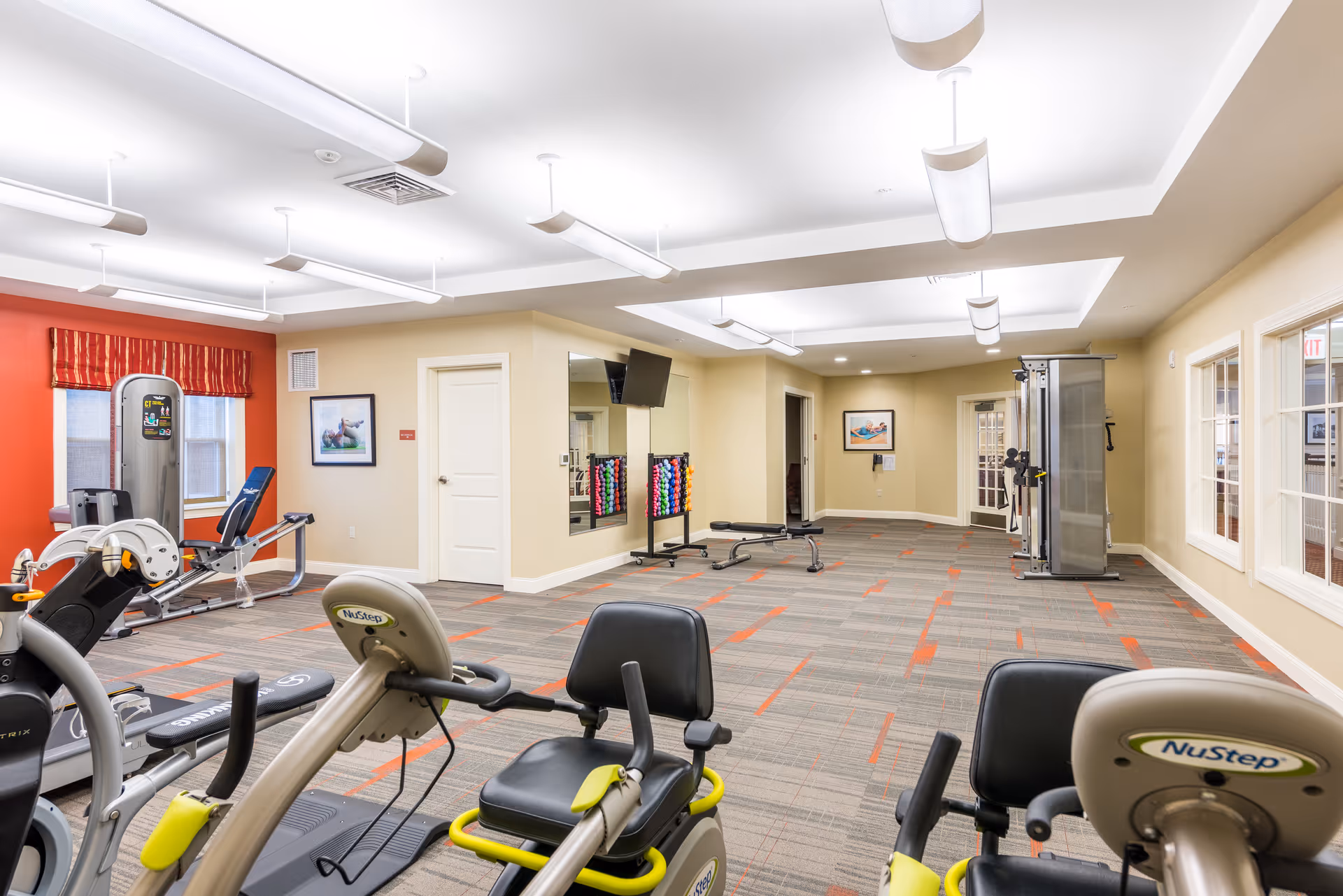 Well-lit fitness room in a senior living facility with exercise machines, weight benches, free weights and wall-mounted TVs.