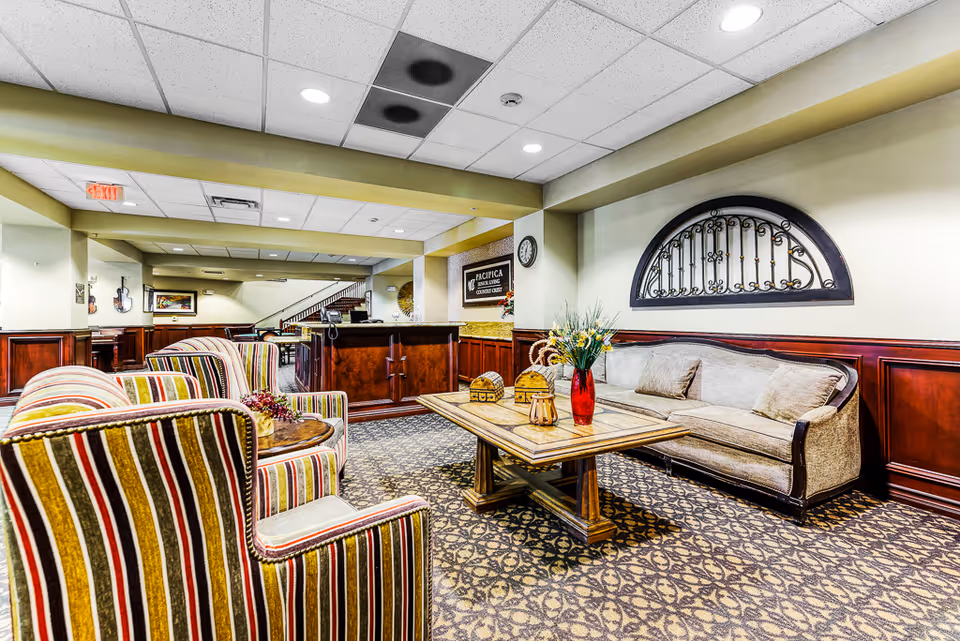 A furnished senior living common area with striped armchairs, a sofa, coffee table, floral arrangement, and a reception desk under ceiling lights.