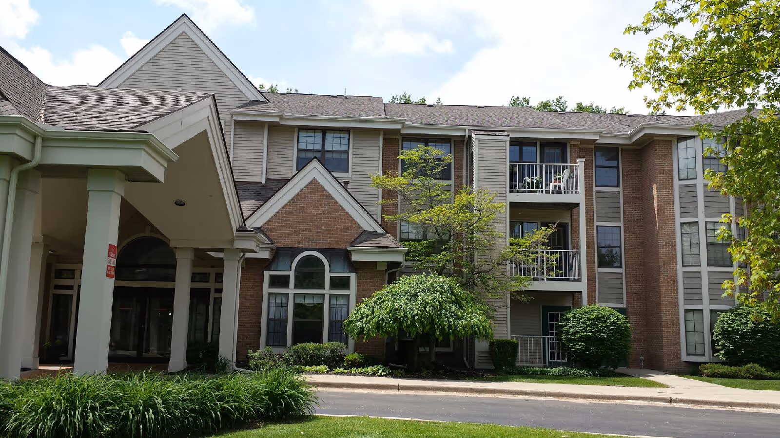 Exterior view of Porthaven Manor, a multi-story senior living facility with beige siding and brick accents. The building features balconies, large windows, and a covered entrance supported by columns. There are well-maintained shrubs and trees in front of the building under a partly cloudy sky.