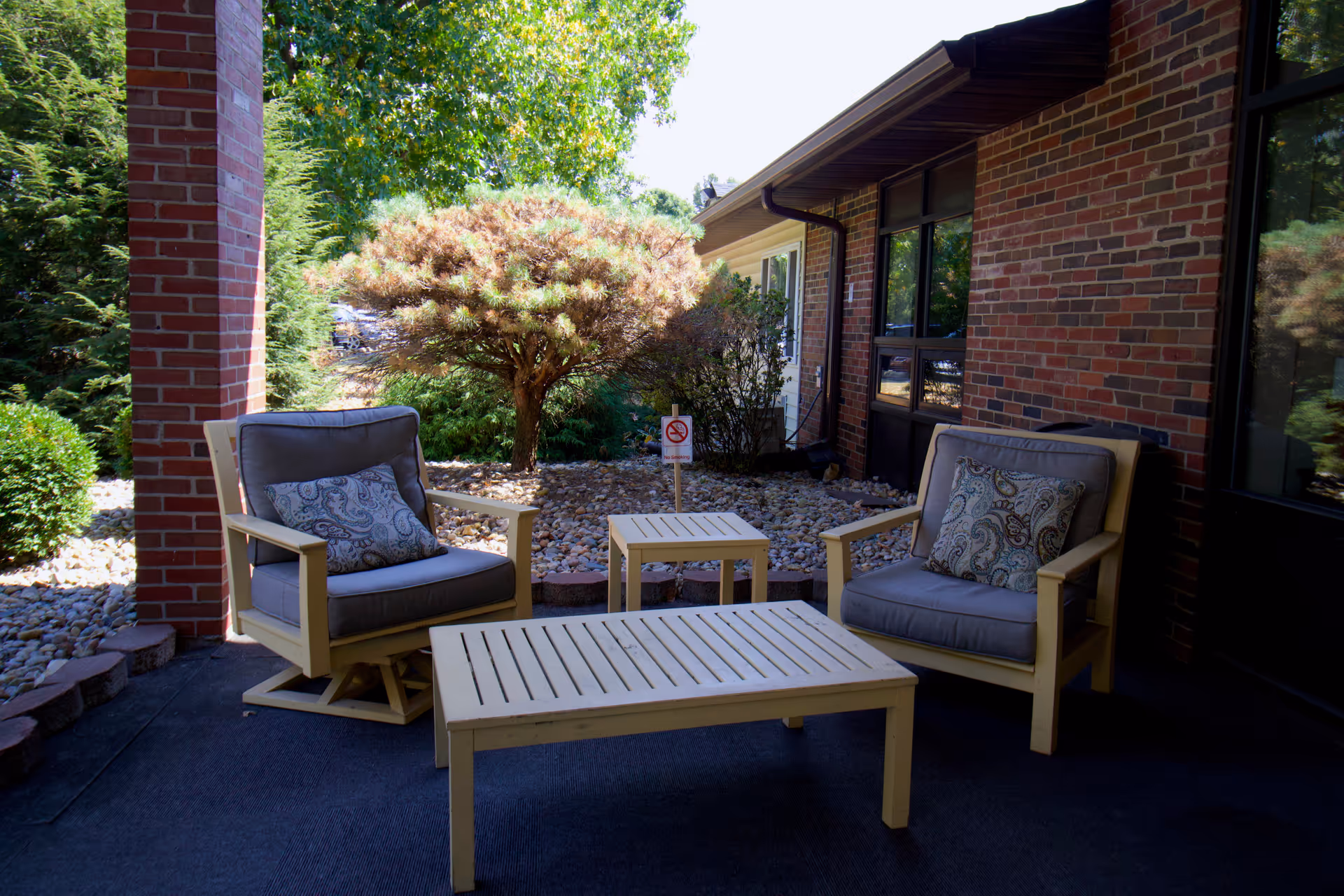 Outdoor patio area with two cushioned armchairs and a matching coffee table and side table. The patio is adjacent to a brick building and surrounded by landscaping with rocks, bushes, and a small tree.