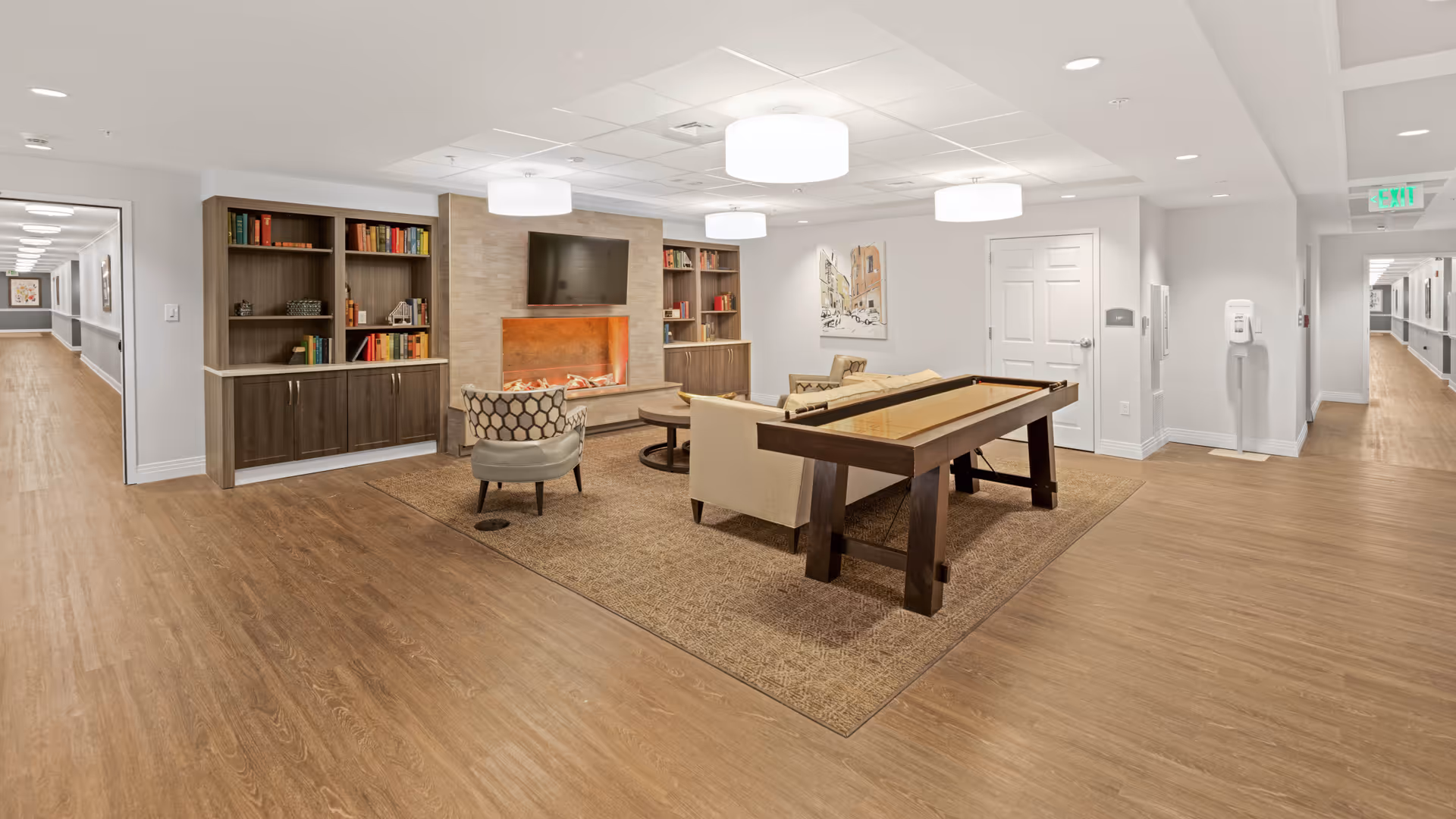 A spacious and well-lit common area in a senior living facility featuring a seating arrangement with a sofa, armchairs, a coffee table, and a shuffleboard table on a large rug. The room has built-in bookshelves filled with books, a wall-mounted TV above a modern electric fireplace, and neutral-colored walls and flooring. Two hallways extend from the room, and ceiling lights illuminate the space.