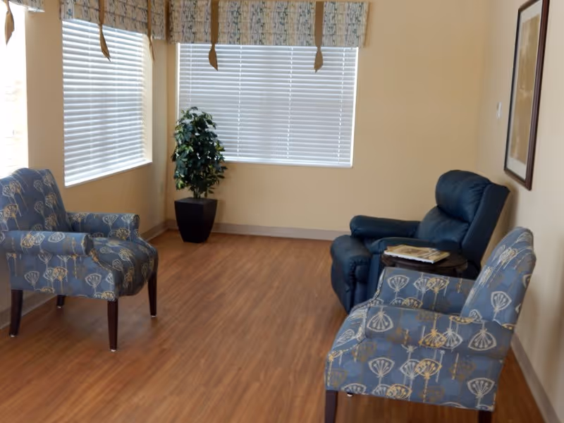 A small sitting area with two patterned armchairs and one blue recliner chair arranged around a small round table with magazines. There are two windows with blinds and valances, a potted plant in the corner, and a framed picture on the wall. The floor is wood laminate.