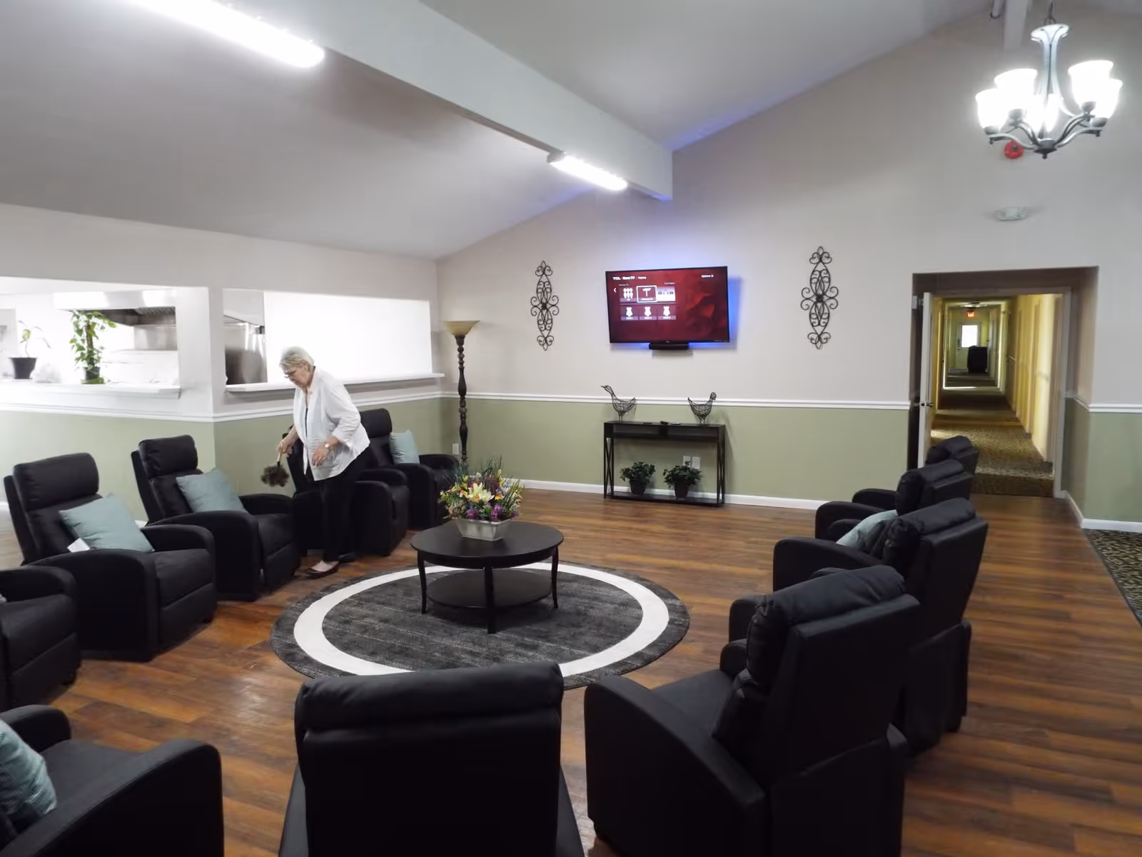 A senior living facility common area with black recliner chairs arranged in a circle around a round coffee table with a flower arrangement. A woman is standing near one of the chairs. The room has wood flooring, a wall-mounted TV, decorative wall hangings, a floor lamp, and a hallway leading to other rooms.