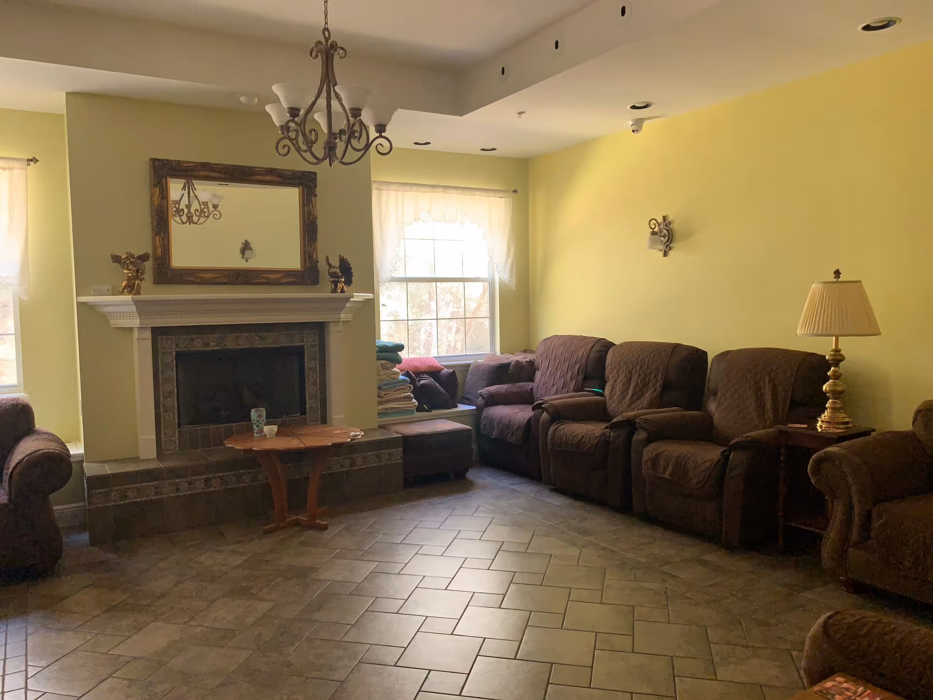 A cozy living room with yellow walls, a tiled floor, and a fireplace with a large framed mirror above it. There are several brown upholstered armchairs and a small wooden coffee table in front of the fireplace. A window with sheer curtains lets in natural light, and a brass table lamp sits on a side table next to one of the armchairs. A chandelier hangs from the ceiling.