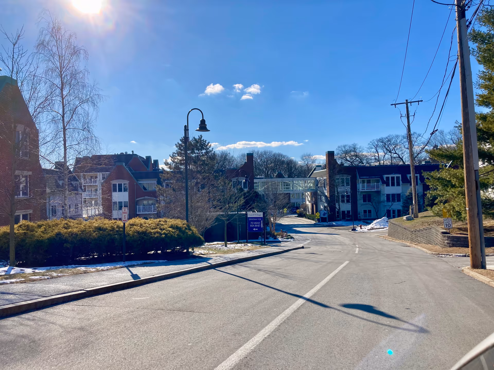 A sunny outdoor view of a senior living facility named Lasell Village, showing a paved road leading to multiple brick and white buildings connected by a glass walkway. There are some leafless trees, bushes, and patches of snow on the ground under a clear blue sky.
