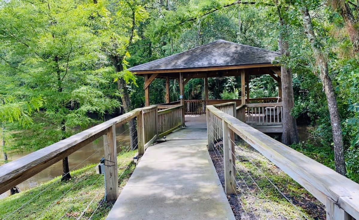 A wooden walkway with railings leads to a covered gazebo surrounded by lush green trees and vegetation near a body of water.