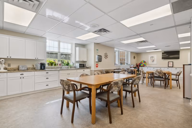 A bright and clean dining area in a senior living facility with wooden tables and cushioned chairs arranged neatly. The room has white cabinets along one wall with a countertop holding kitchen appliances such as a toaster, coffee maker, and microwave. Large windows let in natural light, and the ceiling has recessed lighting panels. There is a wall-mounted TV and a colorful wheel game in the background.