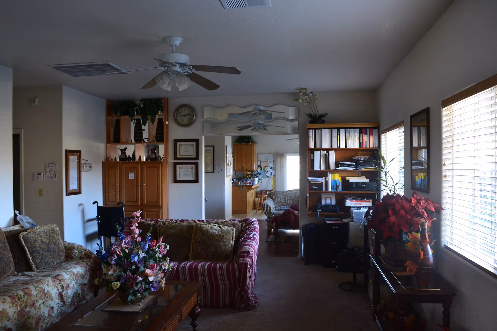 A furnished living room with floral and striped sofas, a coffee table topped with a floral arrangement, bookshelves, a ceiling fan, and window blinds letting in light.