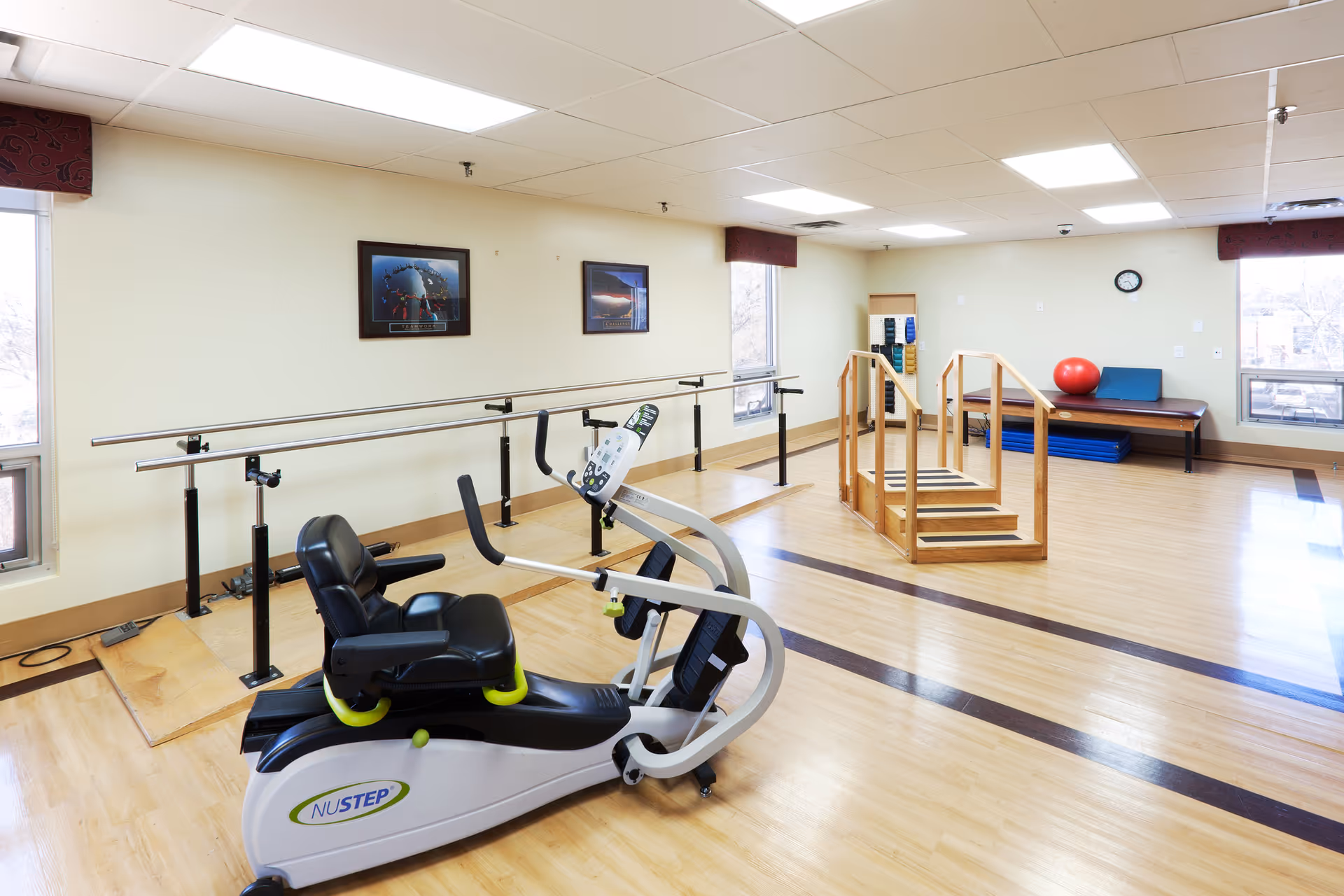 A bright physical therapy or rehabilitation room with wooden flooring, parallel bars for walking exercises, a recumbent exercise bike labeled NUSTEP, a set of wooden stairs for step training, a bench with a red exercise ball and blue mats, and two framed pictures on the wall. Large windows allow natural light into the room.