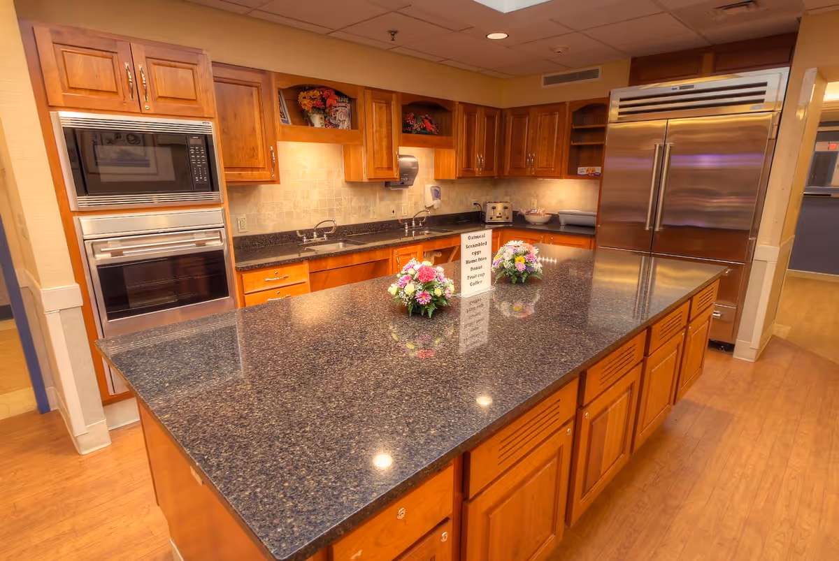 A spacious kitchen with wooden cabinets, a large granite island countertop in the center, two flower arrangements on the island, built-in microwave and oven, double sinks, a stainless steel refrigerator, and a menu sign listing breakfast items.
