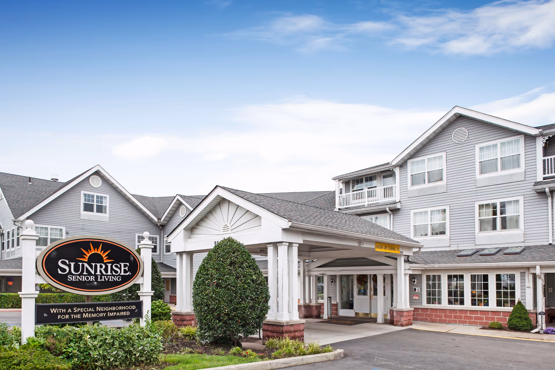 Exterior view of Sunrise Senior Living facility with a covered entrance, manicured bushes, and a clear blue sky.