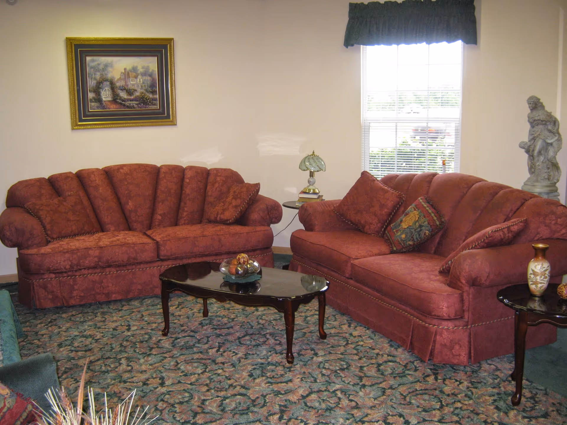 A cozy living room with two red upholstered sofas, a wooden coffee table, patterned carpet, wall art, lamps and a window.