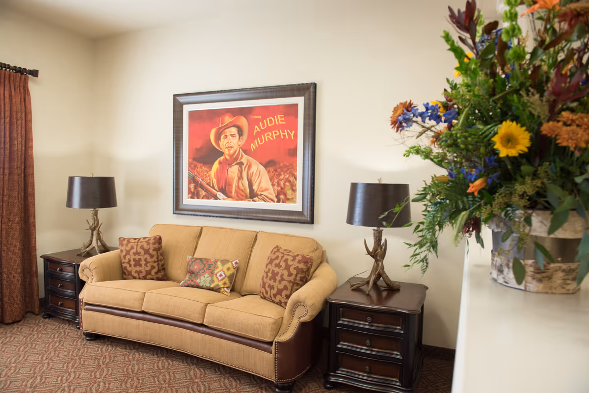 A cozy living room with a beige sofa adorned with three decorative pillows. On either side of the sofa are dark wooden side tables with matching lamps featuring unique branch-like bases. Above the sofa hangs a framed vintage-style poster of Audie Murphy. To the right, there is a large floral arrangement with various colorful flowers in a rustic pot. The room has beige walls, a patterned carpet, and a brown curtain on the left side.