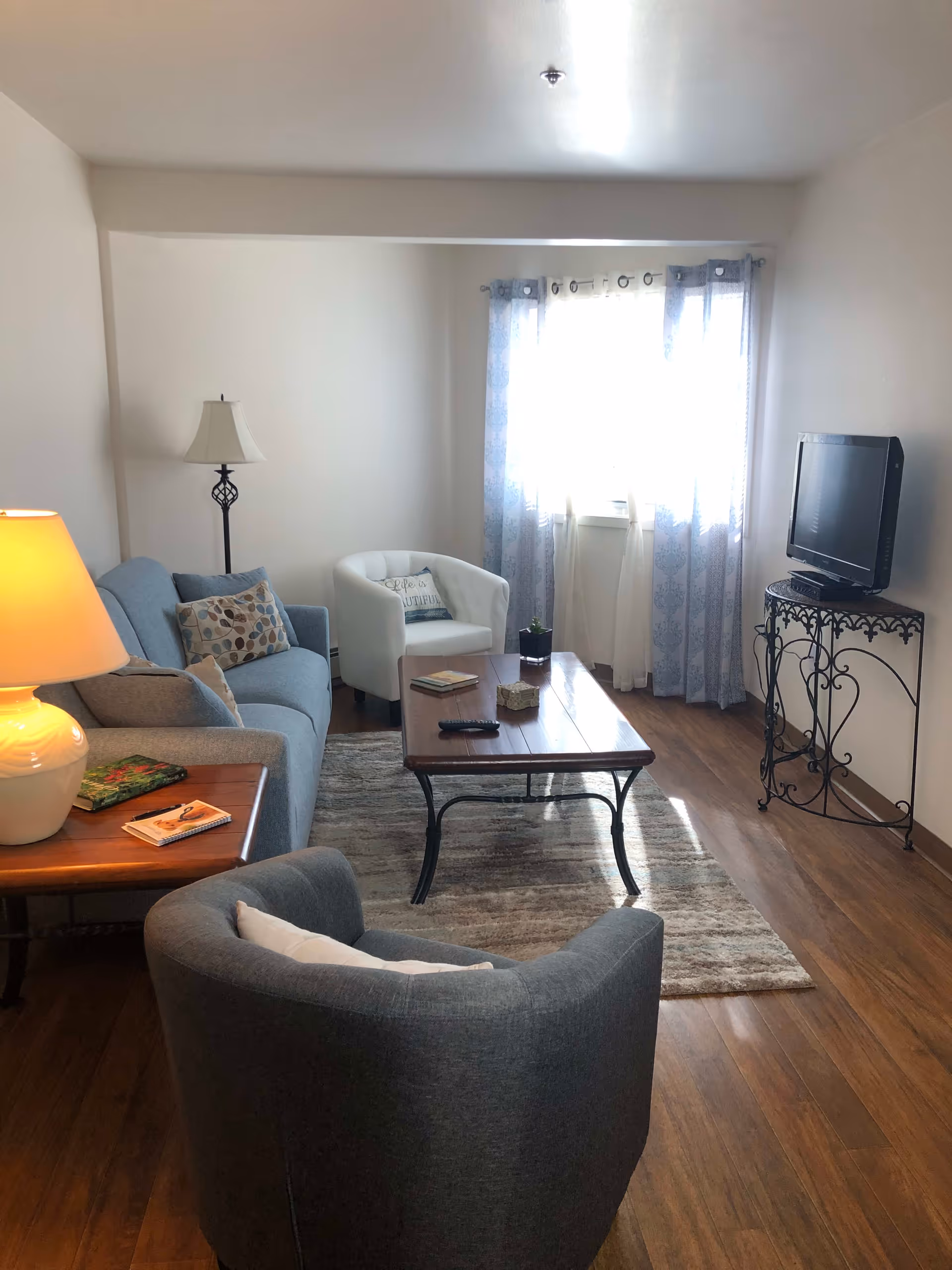 A cozy living room with a gray couch, a white armchair, and a dark gray armchair arranged around a wooden coffee table on a patterned rug. There is a floor lamp next to the couch and a table lamp on a side table with books. A TV is placed on a decorative metal stand near a window with sheer and patterned curtains, letting in natural light. The room has wooden flooring and white walls.