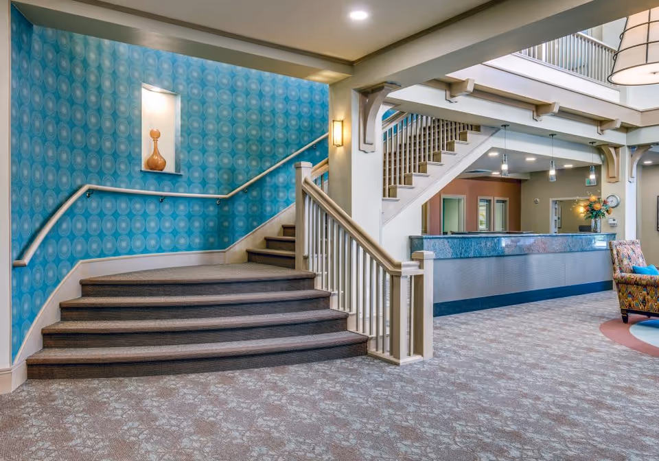 Interior view of a senior living facility lobby with a carpeted staircase featuring a blue patterned wall on the left. The lobby includes a reception desk with a blue countertop, pendant lights hanging from the ceiling, and a colorful upholstered chair on the right. The area is well-lit with ceiling lights and has a spacious, welcoming atmosphere.