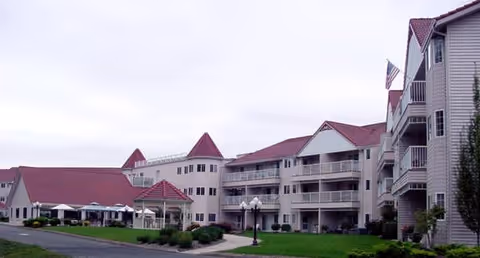 Exterior view of Riverview Terrace senior living facility showing multiple connected buildings with red roofs, balconies, and a well-maintained lawn with shrubs and a walkway.