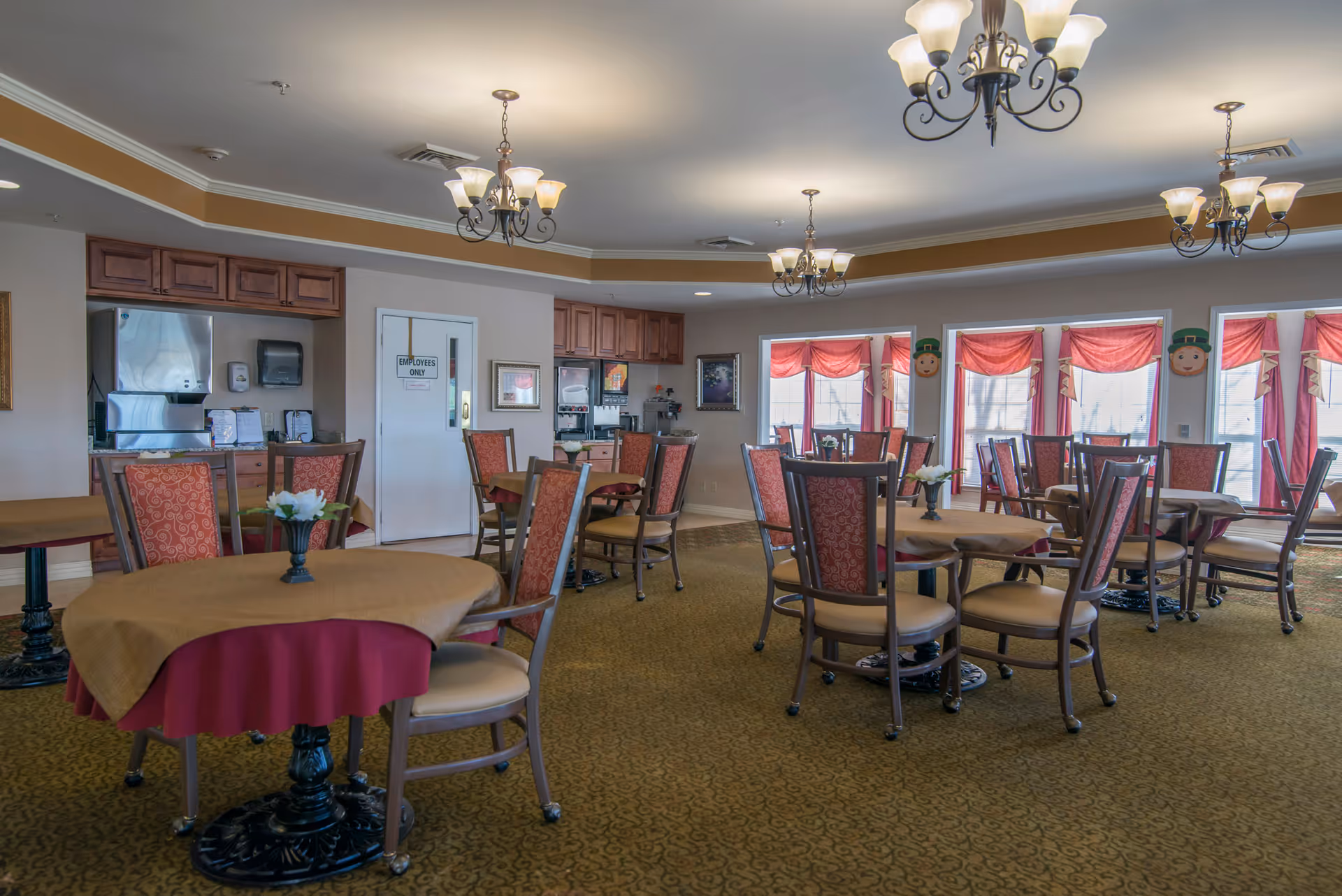 A spacious dining room with multiple round tables covered with beige and burgundy tablecloths. Each table is surrounded by wooden chairs with red patterned cushions. The room features several chandeliers hanging from the ceiling, large windows with red valances, and a carpeted floor. There is a counter area with cabinets and a door labeled 'Employees Only' in the background.