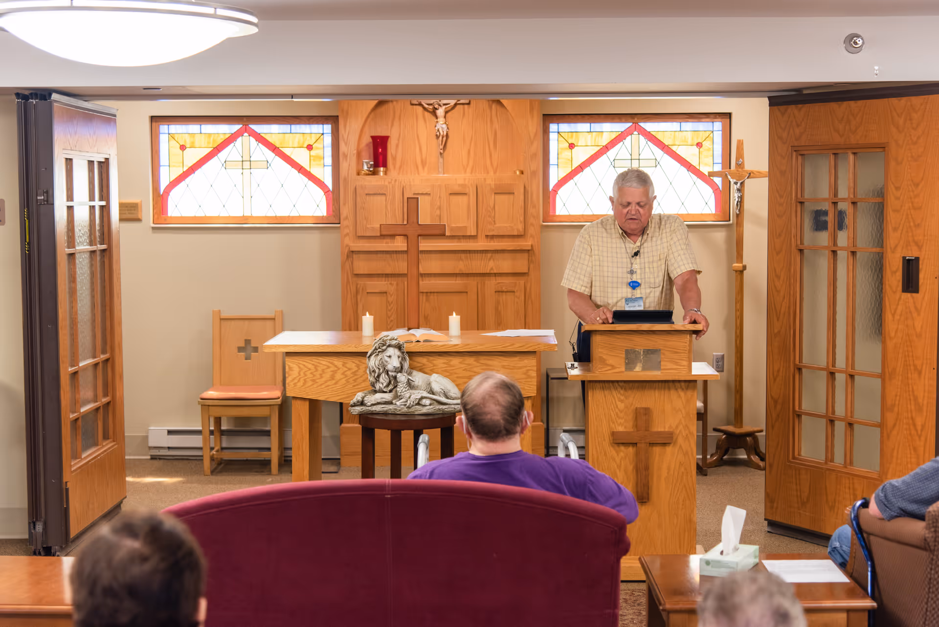 A man stands at a wooden lectern in a small chapel speaking to seated residents.
