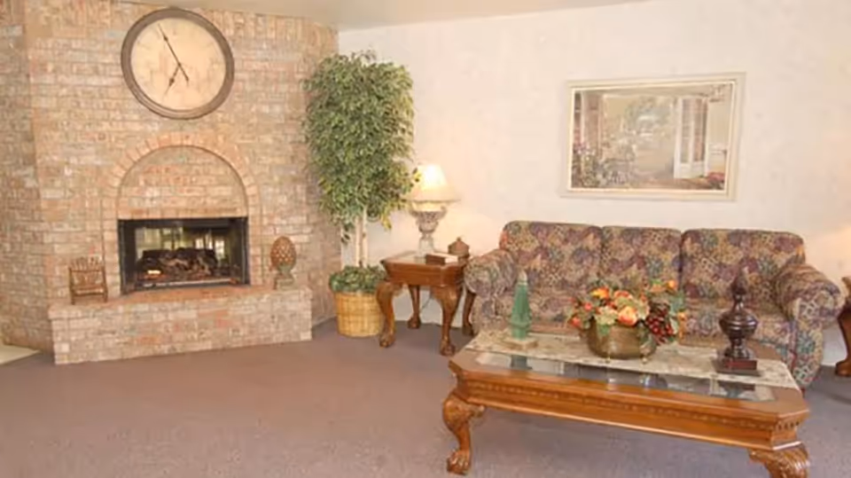 Cozy interior lounge with a brick fireplace, patterned sofa, coffee table, side table with lamp, and a potted plant.