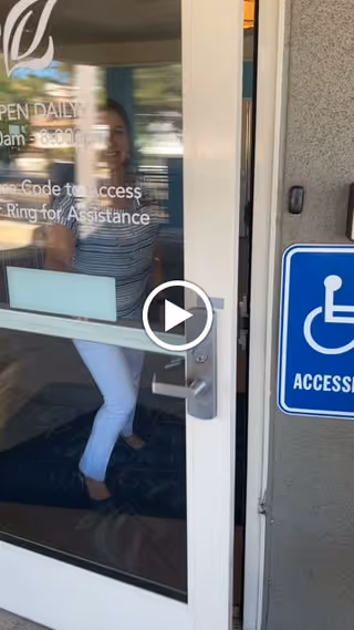 A woman wearing a striped shirt and white pants is seen through a glass door that is partially open. The door has text indicating daily open hours and instructions for access. A blue accessibility sign with a wheelchair symbol is mounted on the wall next to the door.