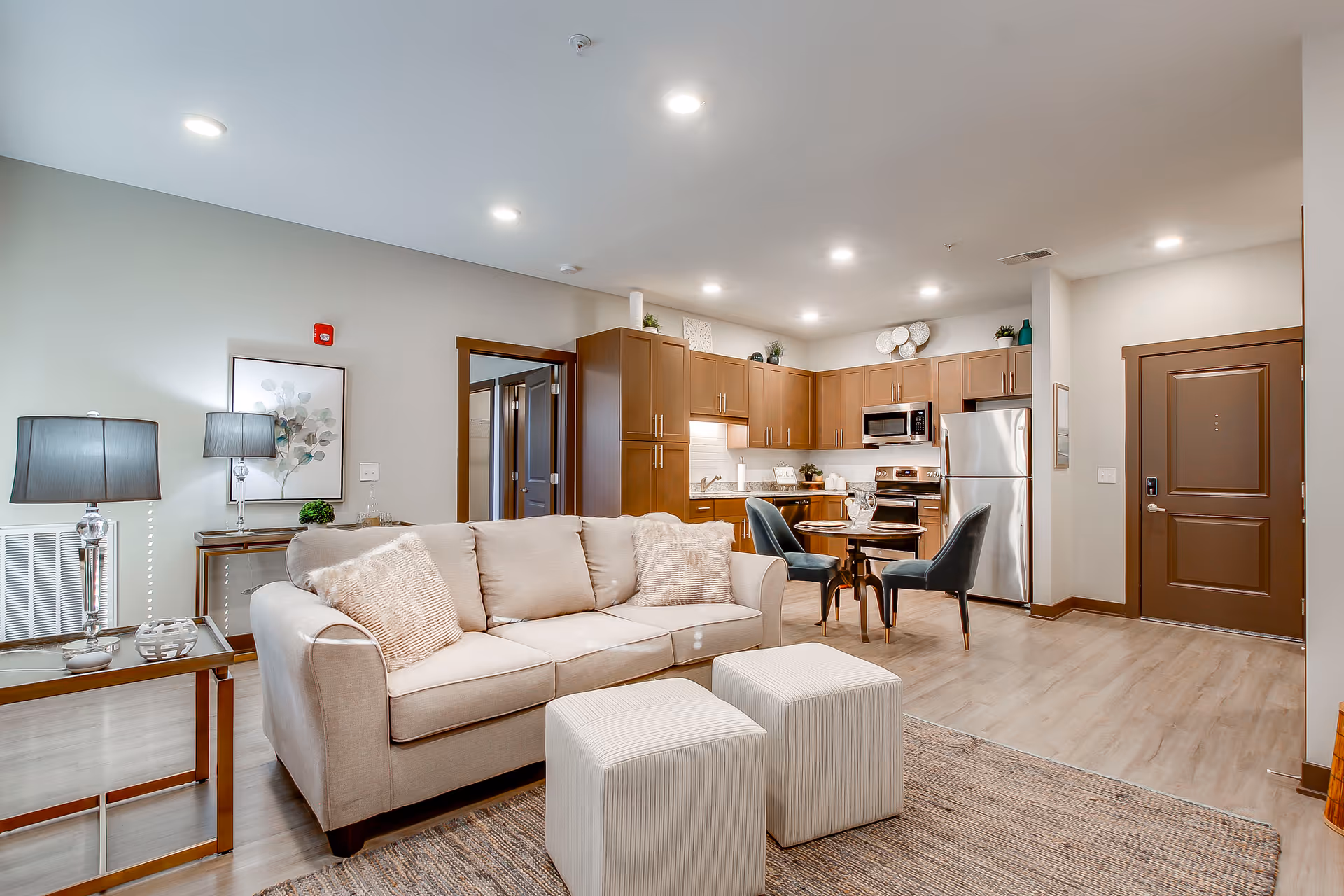 A modern open-concept living space featuring a beige sofa with two matching ottomans on a textured rug. Behind the sofa is a small table with a lamp and decorative items. The kitchen area includes wooden cabinets, stainless steel appliances, and a small round dining table with two blue upholstered chairs. The room has light wood flooring and recessed ceiling lights.