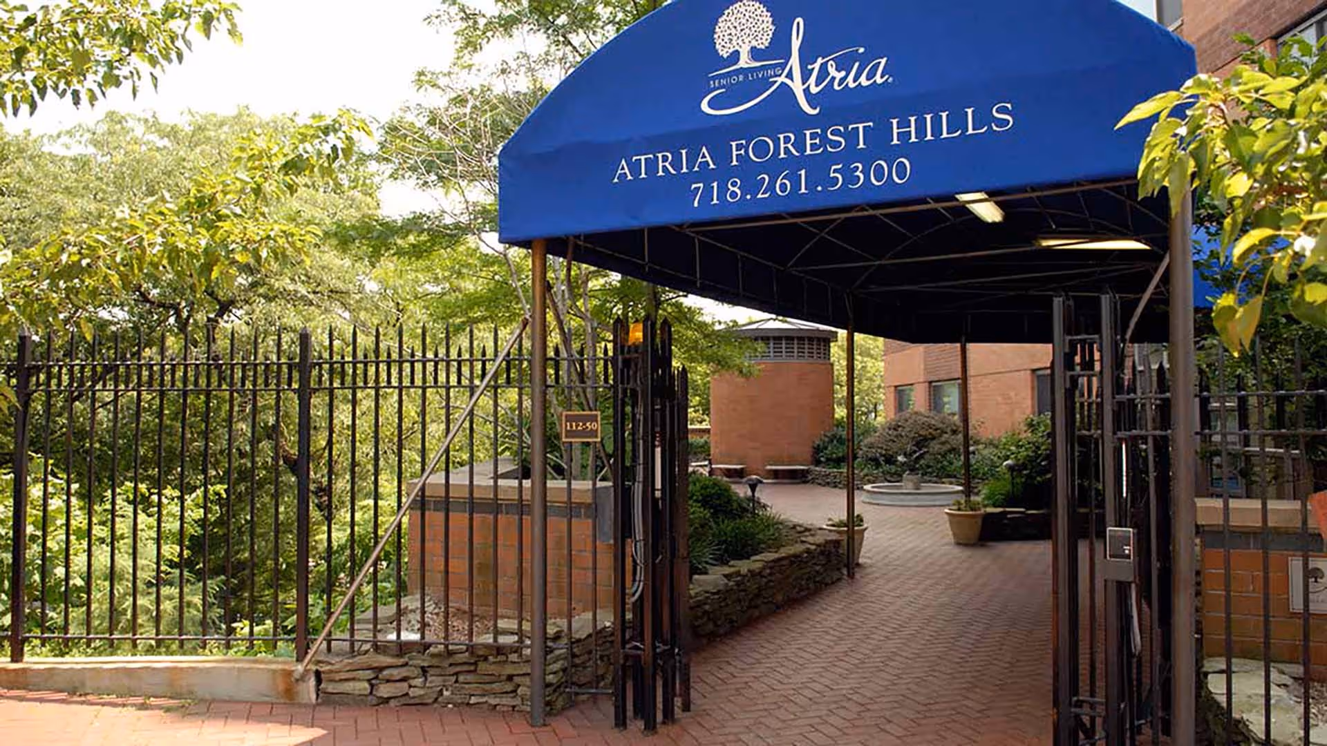 Entrance to Atria Forest Hills senior living facility with a blue canopy displaying the facility name and phone number, surrounded by greenery and a brick pathway leading inside.