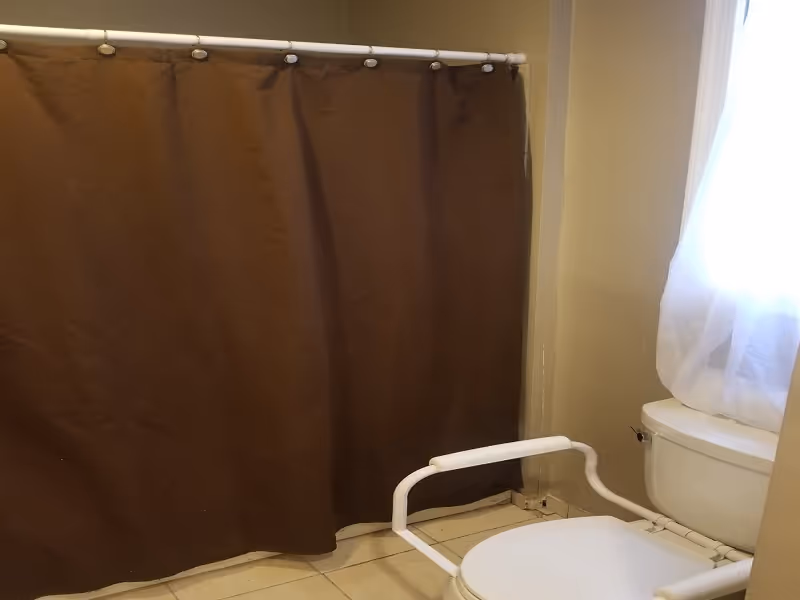 Bathroom with a brown shower curtain, a white toilet with safety rails, and a window with a sheer white curtain allowing natural light to enter.