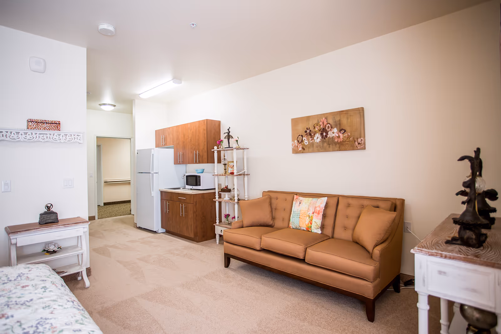 Well-lit studio living area with a tan sofa, wall art, and a small kitchenette with refrigerator and cabinets.